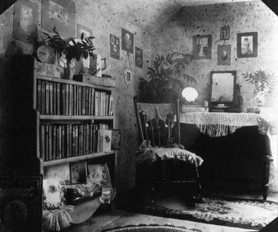 Black-and-white photograph of a room, a filled bookcase on the left, a chair in the centre, and a table with a mirror on the right. There are pictures hung on the wall.  