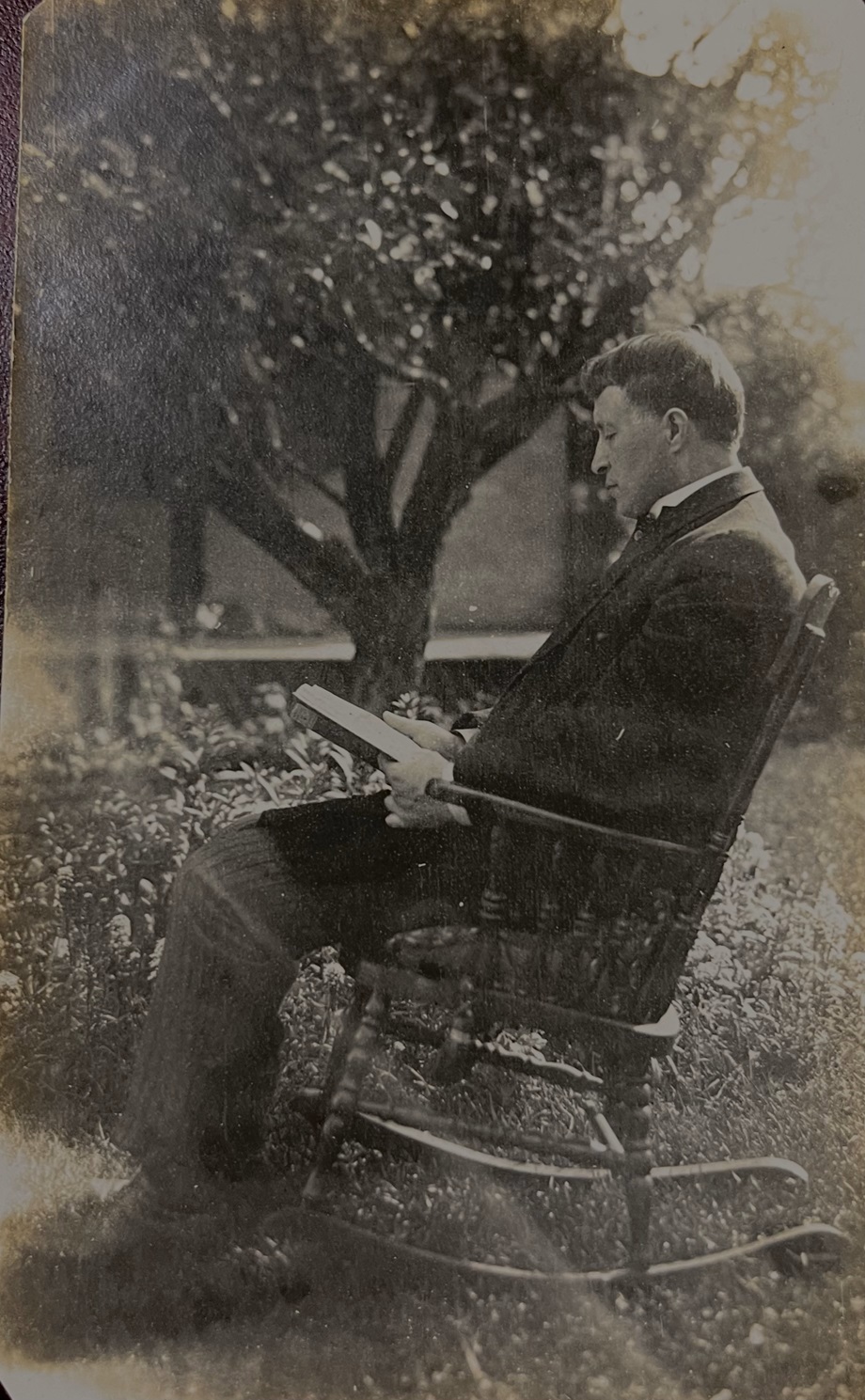A side-view of Ewen Macdonald, with dark hair, in a rocking chair on the lawn beside a tree and a house. He is reading a book and wearing a suit.
