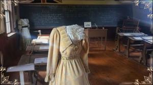 A human-sized mannequin without a head in Victorian clothing standing in front of a school desk in an old classroom.