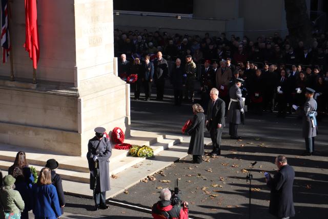 Andrea McKenzie A broader view of the Cenotaph. Andrea stands with veteran soldiers behind her. A soldier stands with bowed head at each corner of the monument. School children are standing at the left. Veterans, their families, and other spectators stand on the right.