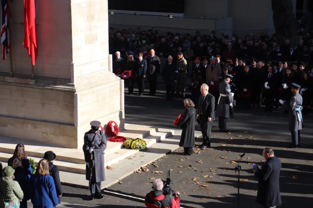 A broader view of the Cenotaph. Andrea stands with veteran soldiers behind her. A soldier stands with bowed head at each corner of the monument. School children are standing at the left. Veterans, their families, and other spectators stand on the right.