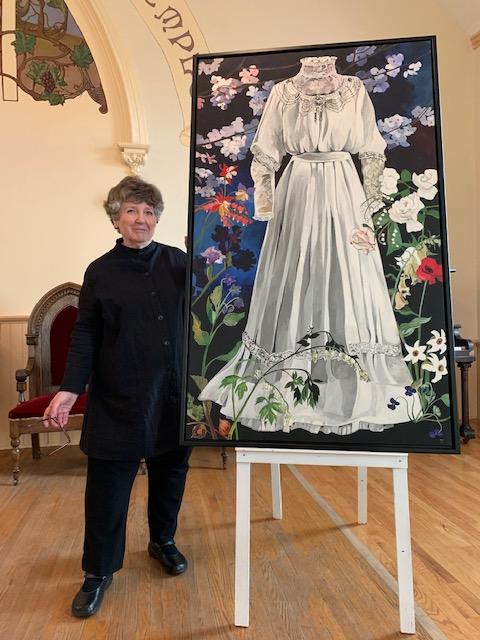 The smiling artist stands to the left of a painting of a white wedding dress surrounded by colourful flowers including white roses at the waist.