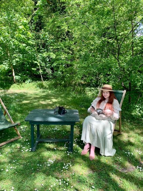 Chelsea Wallis wearing a straw hat and reading a book while sitting in a sunny garden.
