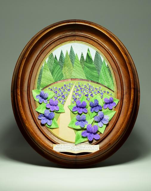 A sculpted paper image of purple flowers with a background of evergreen trees, displayed in a round wooden frame.