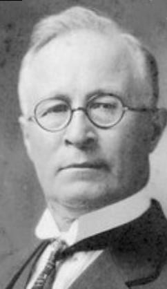 A formal headshot portrait of Ewen Macdonald, as an older man, with grey hair wearing glasses.