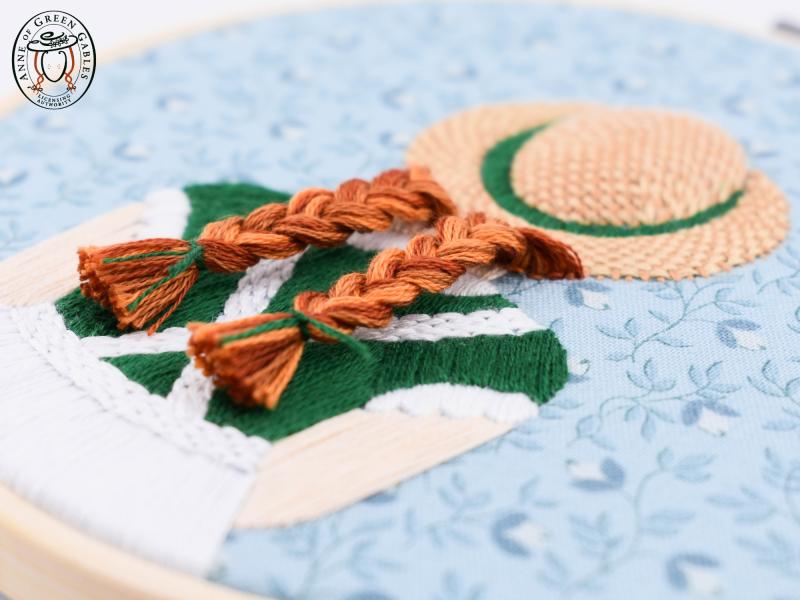 Embroidery depicting red braids under a straw hat.