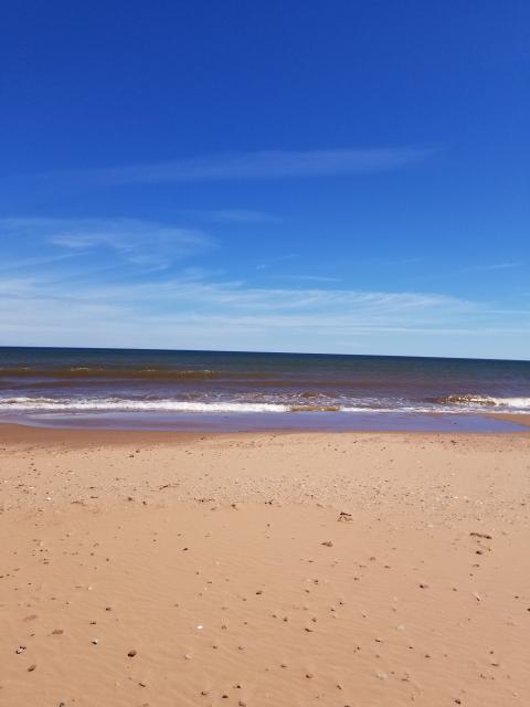 A photograph of a shoreline with small waves rolling onto the red sand. The horizon merges with faint white clouds under a deep blue sky.