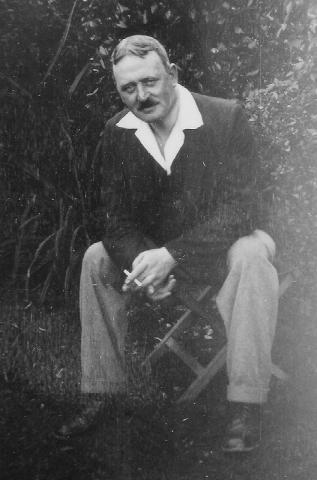 Photograph of George MacMillan sitting on a bench outdoors holding a cigarette. 