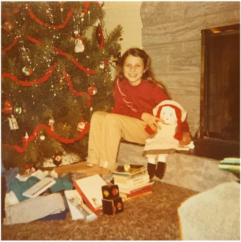 A child sits in front of a Christmas tree near an indoor fireplace.