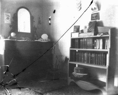 Black-and-white photograph of a room with a vanity on the left and a bookcase on the right. There are several crack lines in the bottom left-hand corner of the image. Two of the cracks continue across the image. 