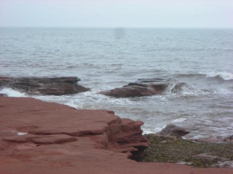 A photograph, taken from a cliff of red rocks, of waves and roiling water crashing over two rocks that are almost submerged.
