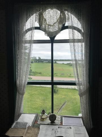 Photo of a lace-curtained framed window overlooking a bay with an oil lamp and quill on the desk in front of it.