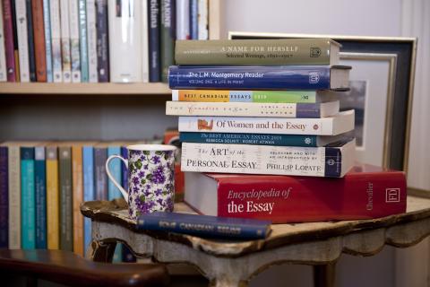 Photograph of a stack of books on an antique table. A teacup with purple flowers is sitting on the table beside the books. There is a filled bookcase in the background.  