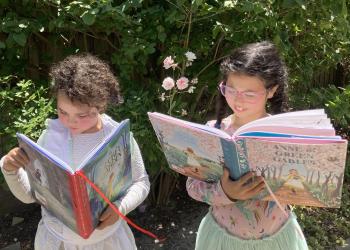 Two young girls reading picture books while standing in a garden.